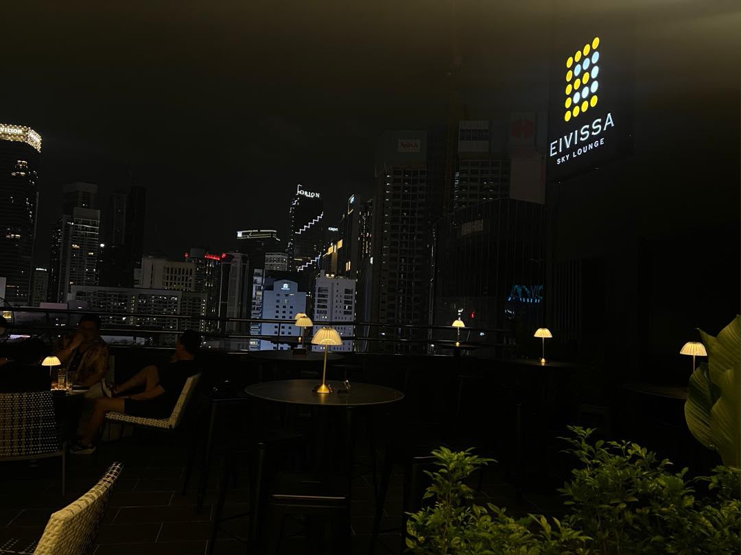 Eivissa Sky Lounge — the rooftop terrace at night, table lamps lit beneath the sign, the Kuala Lumpur skyline beyond the rail.
